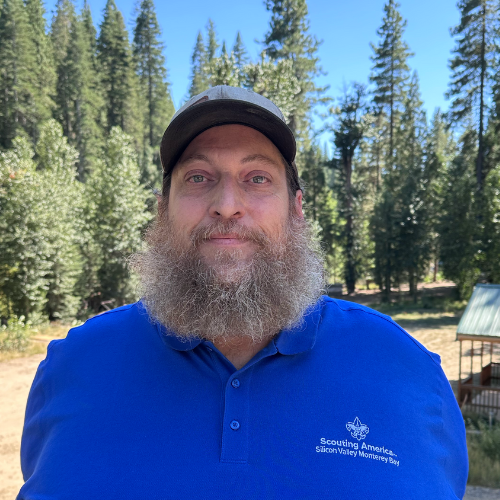 A man in a blue "Scouting America" shirt with trees in the background.